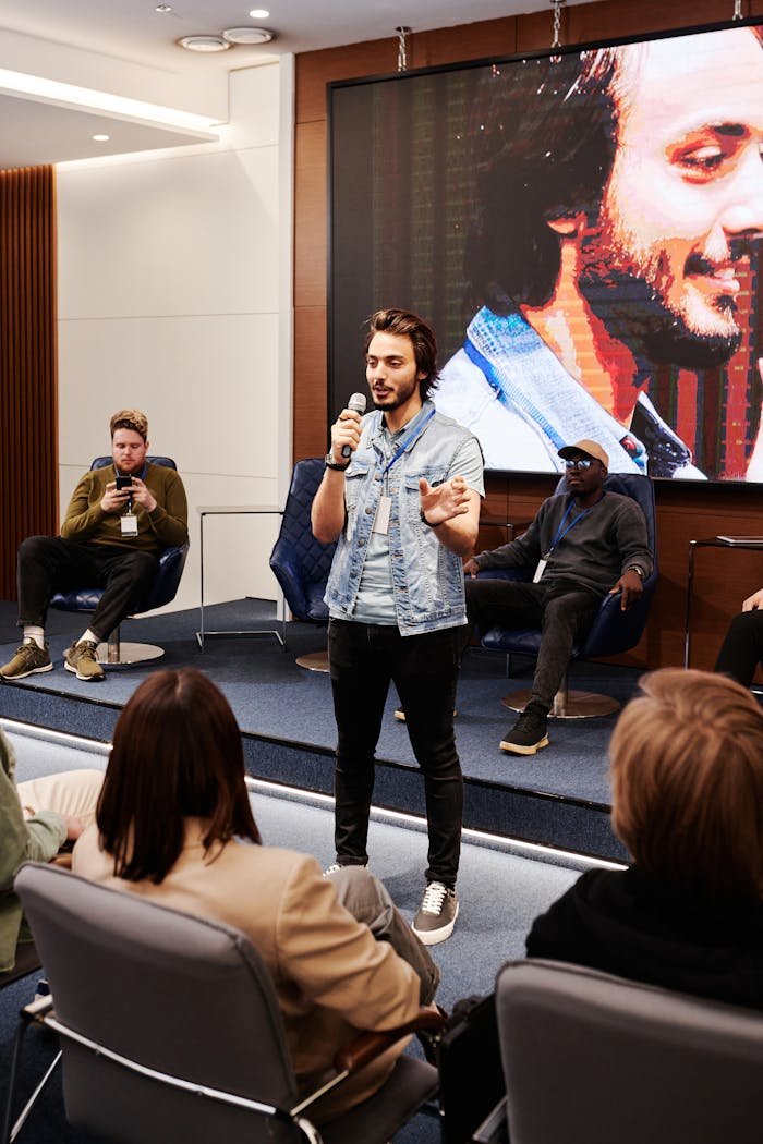 A man holding a microphone speaks at a conference with an audience, in front of a large screen.